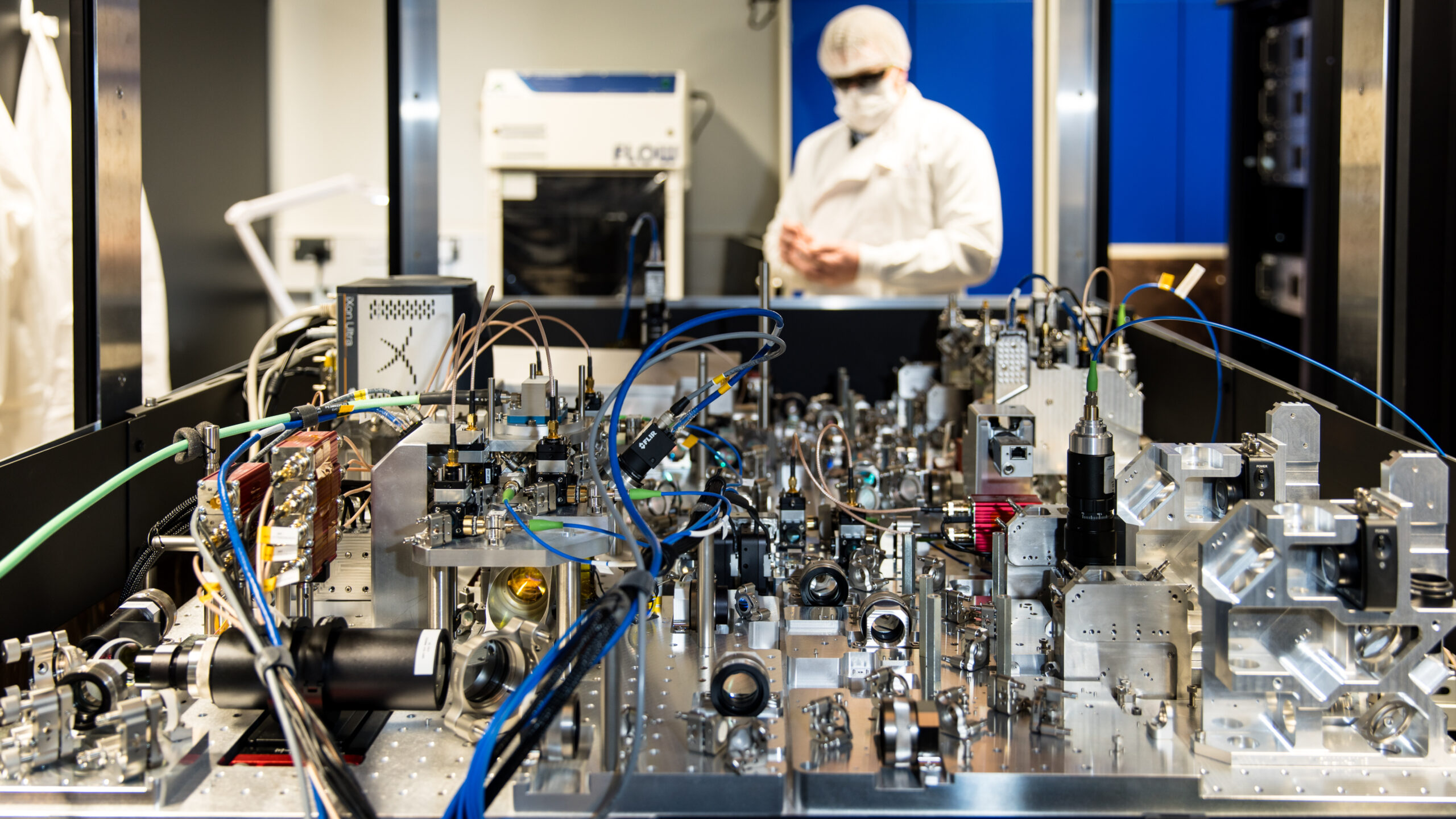 An Infleqtion worker in protective clothing behind an optical laboratory setup