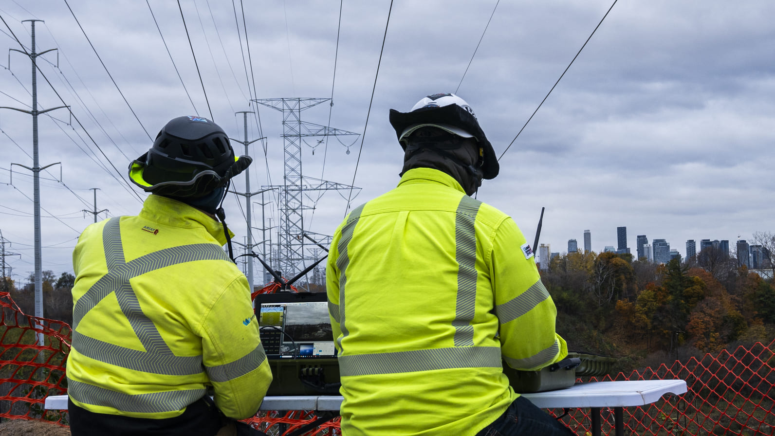 Two field operators in fluorescent safety clothing work at a control station beneath transmission lines