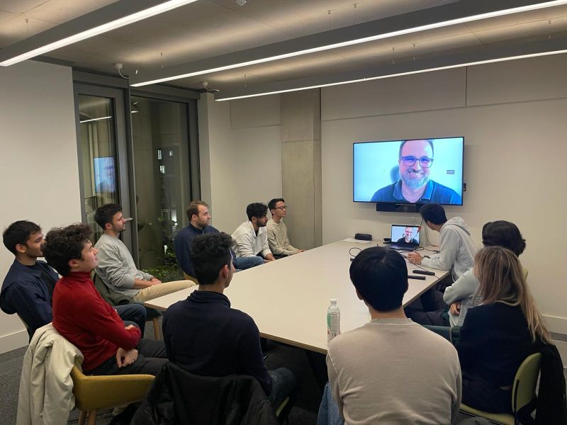 Participants sit around a meeting table during an Initiator community session with a remote speaker on screen