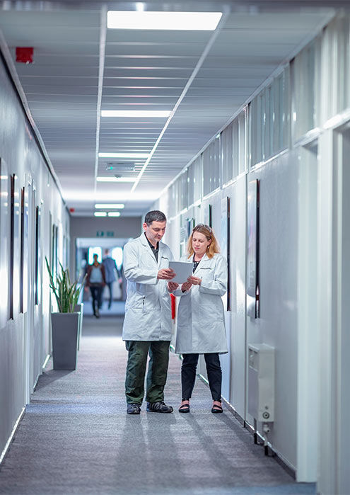 Two colleagues in laboratory coats review a tablet in a corridor