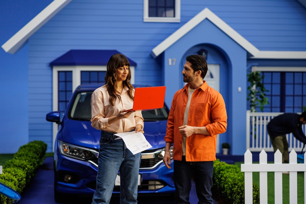 Two actors compare insurance beside a blue car in an Insurify commercial