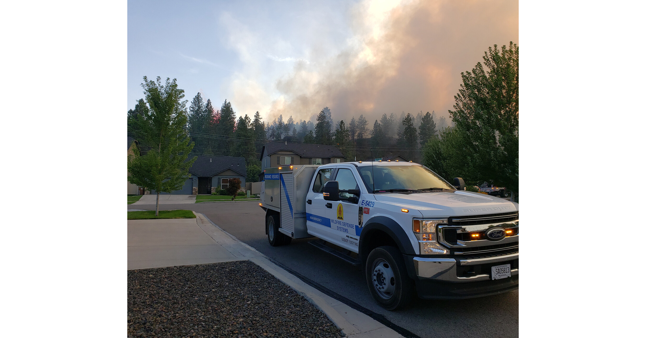 A Wildfire Defense Systems truck near homes under a smoke-filled sky