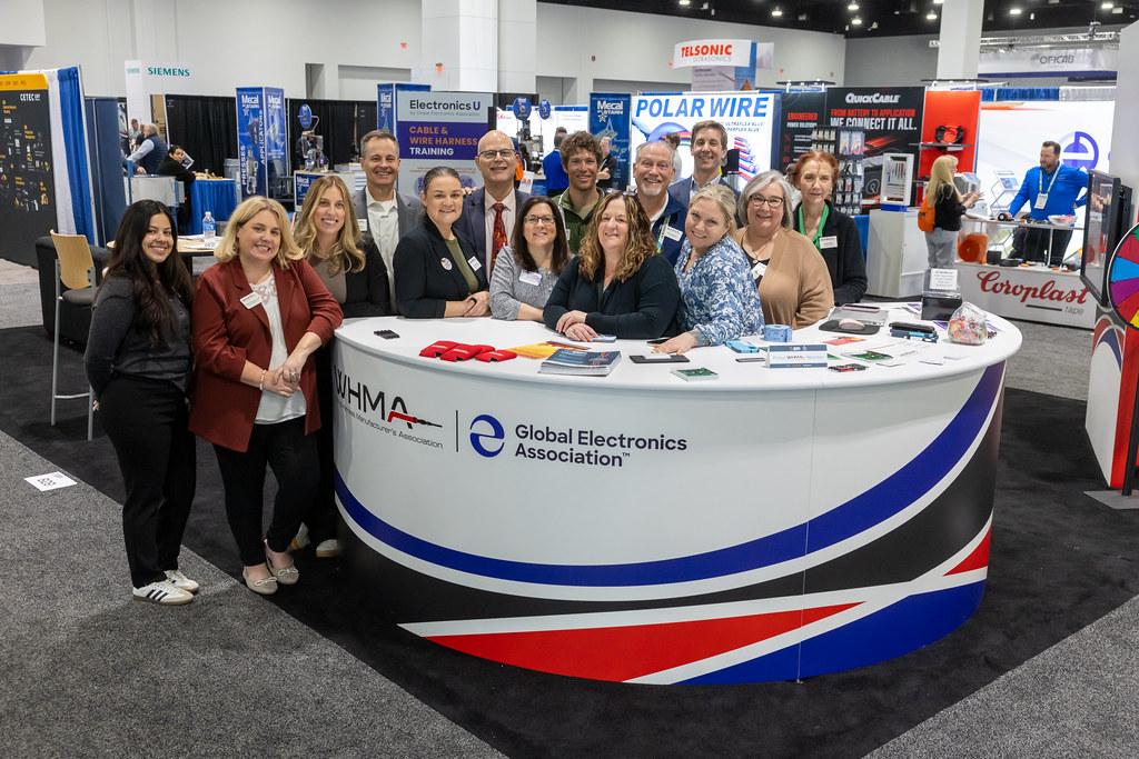 A group gathers at the WHMA and Global Electronics Association booth on an exhibition floor.