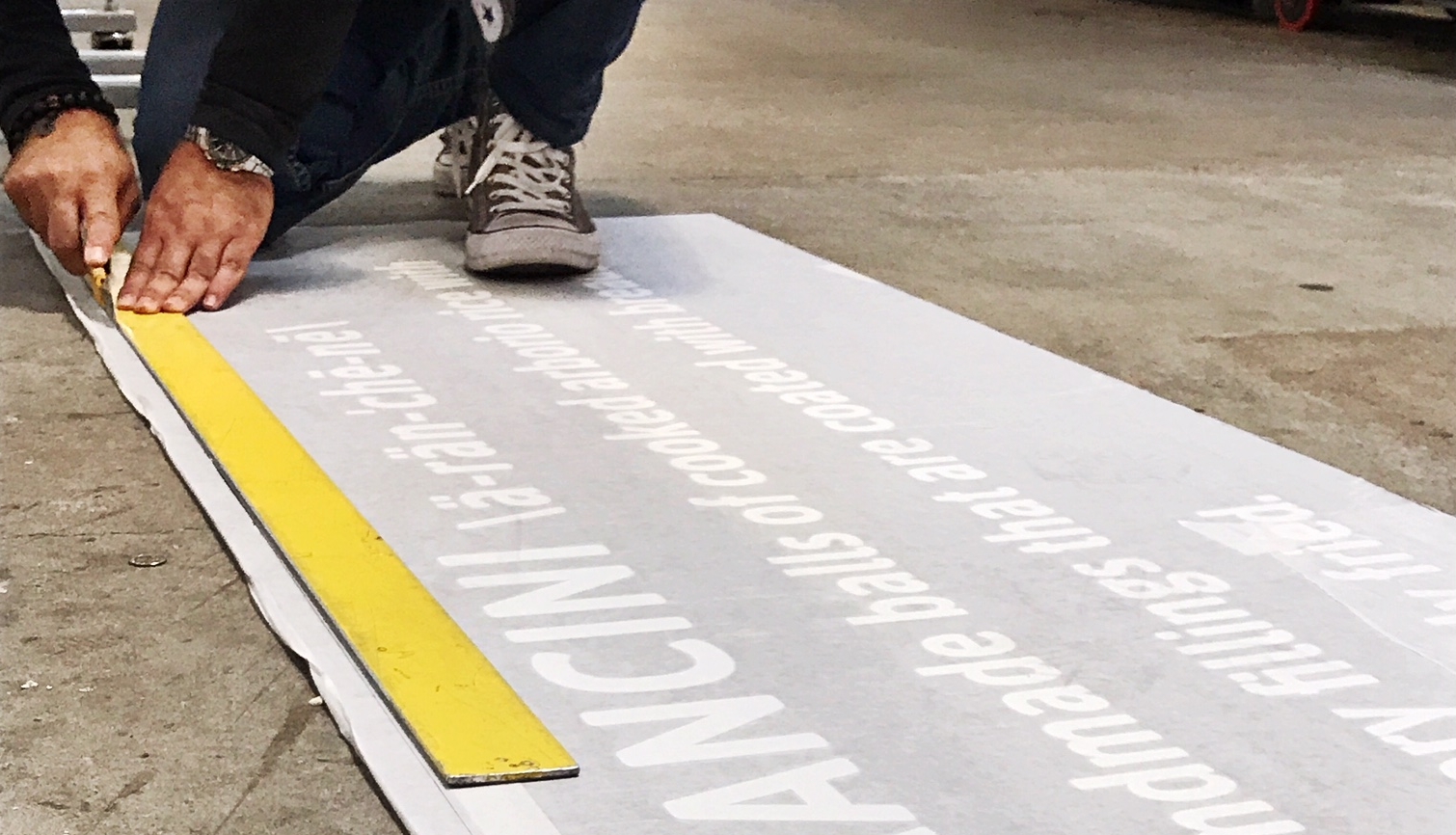 Hands using a yellow straightedge to trim a large printed sign on the floor