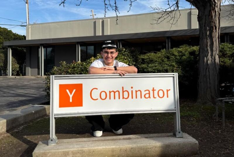 Iqbol Temirkhojaev smiling behind the Y Combinator sign outdoors
