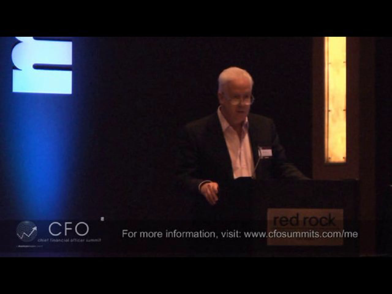 Archival video frame of Ira Birns speaking from a lectern at a CFO summit