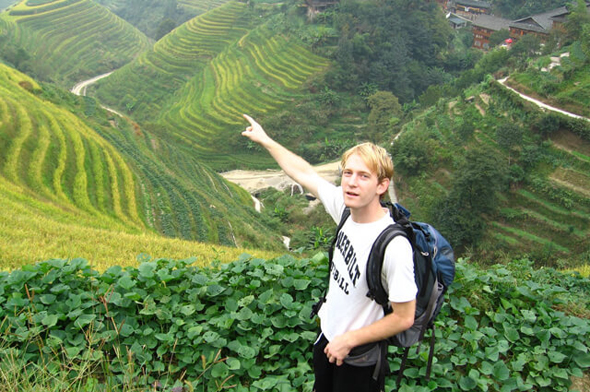 A young Isaac Heller with a backpack, pointing toward terraced fields during his time in China