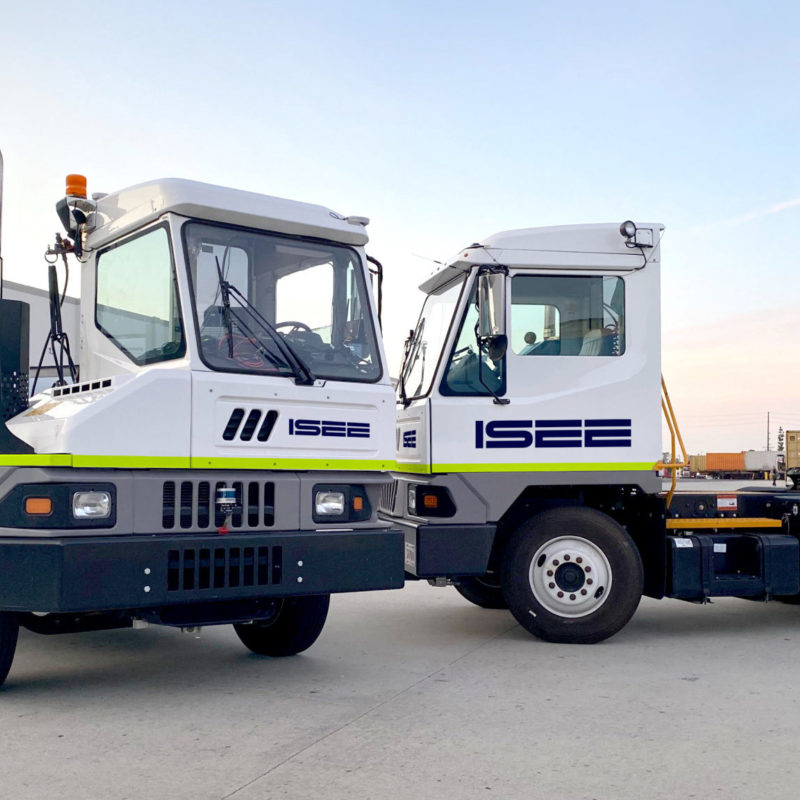 Two ISEE autonomous yard trucks with neon green stripes parked near trailers at dusk