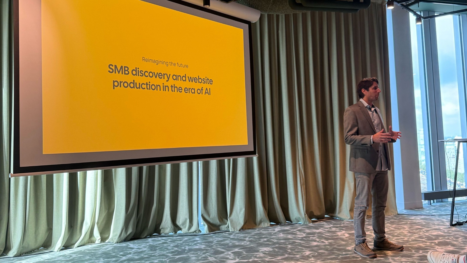 Itai Sadan presenting beside an orange slide about small-business discovery and website production in the AI era