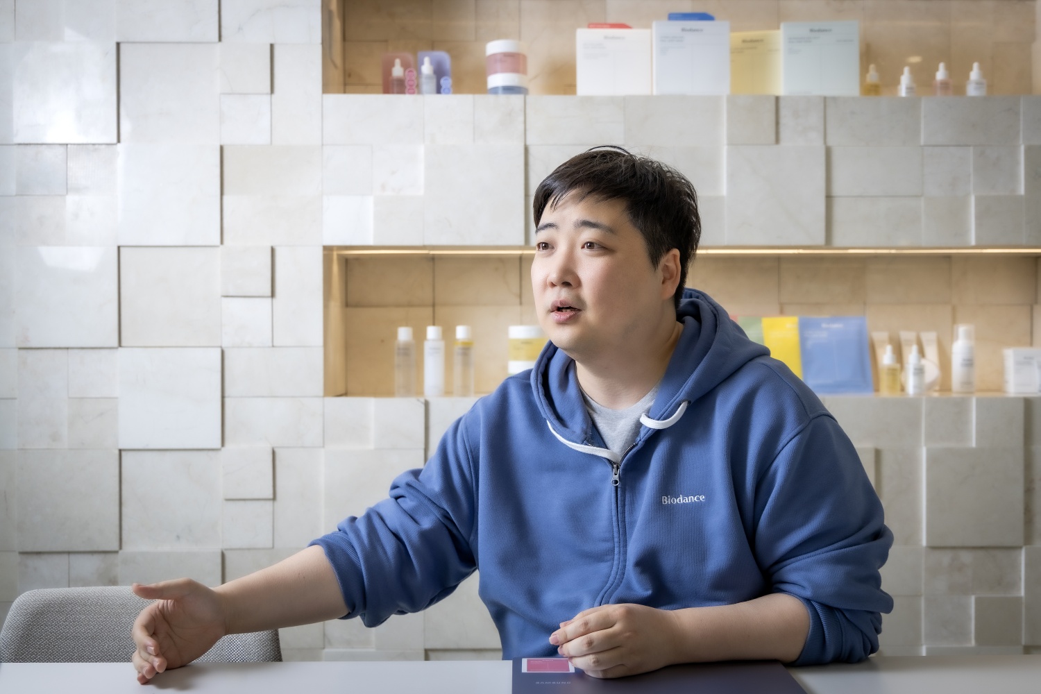 Jae Bin Park in a blue hoodie, speaking at a table with beauty products behind him