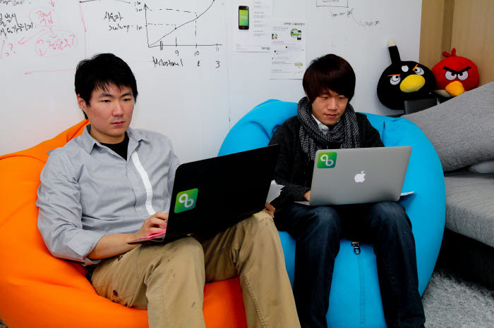 Choi and Kim working on laptops in orange and blue beanbag chairs, with diagrams on the wall