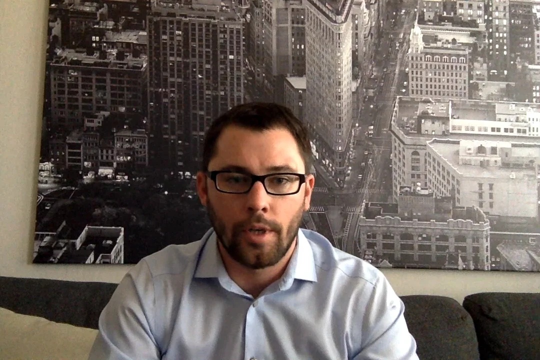 Jake Benson seated on a sofa during a Libra video interview, with a city photograph behind him