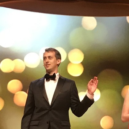 Jake Guglin speaking onstage in a dark suit and bow tie