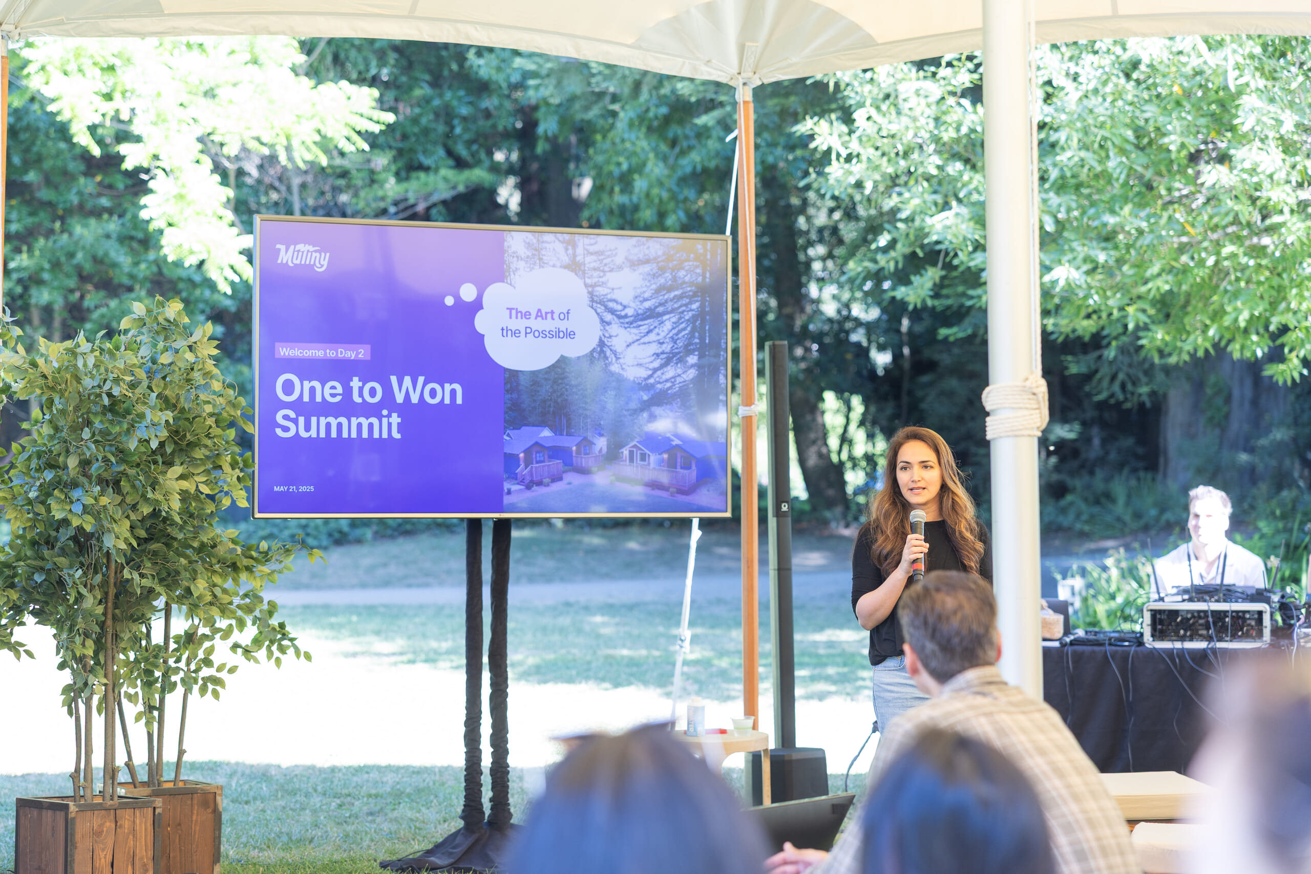 Jaleh Rezaei speaking with a microphone beside a One to Won Summit presentation, outdoors under a canopy
