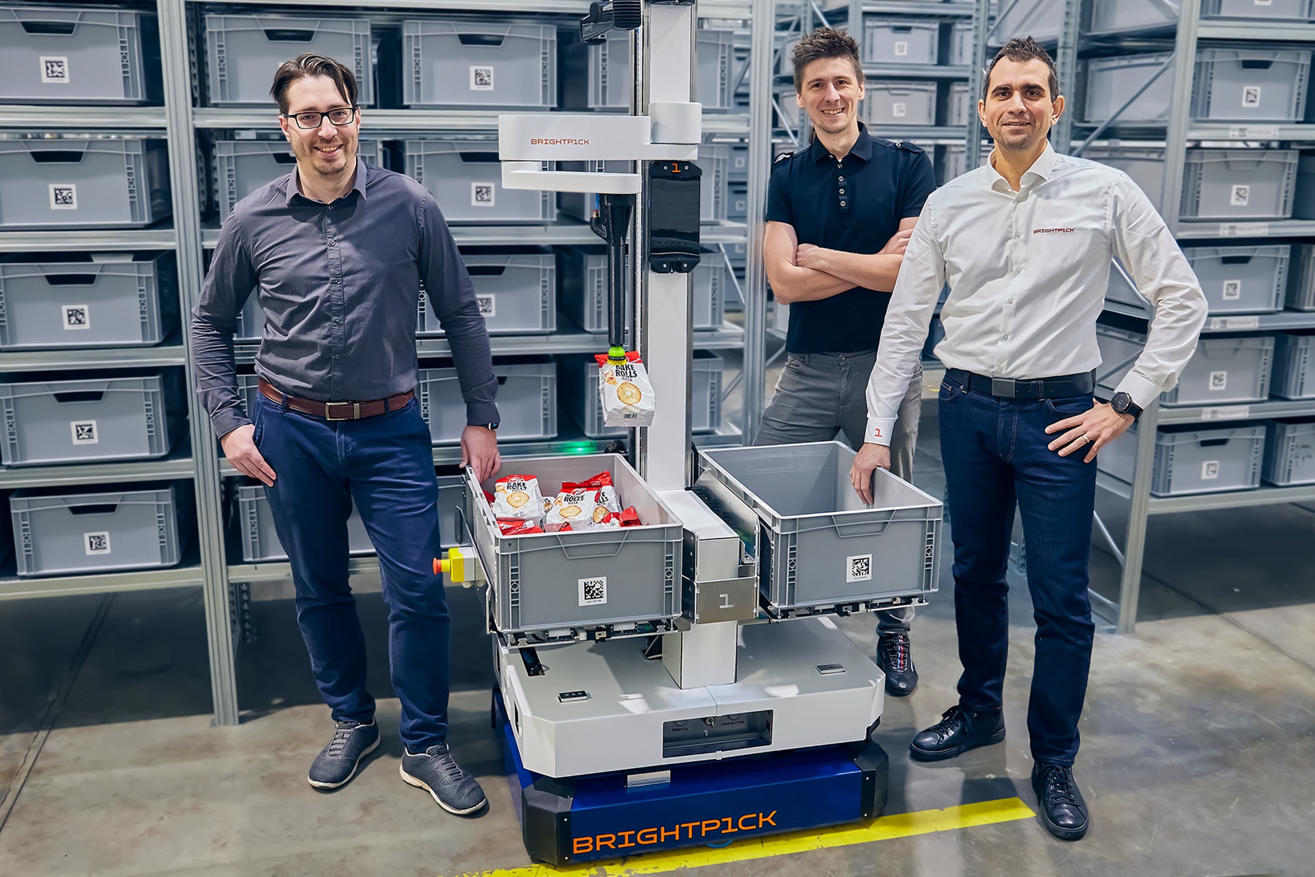 Tomas Kovacovsky, Jan Zizka and Branislav Pulis standing around a Brightpick Autopicker holding a packet of snacks