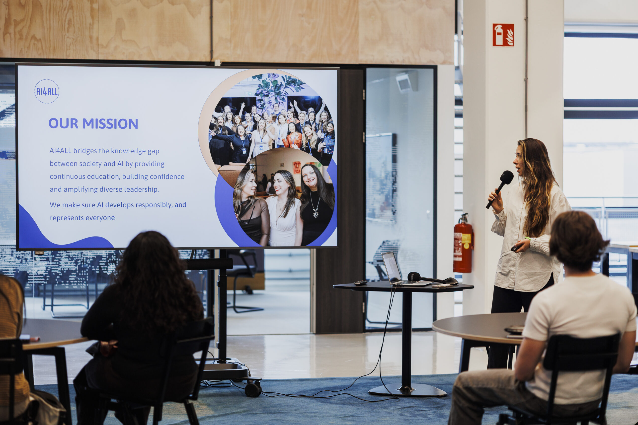 Jana Wilhelmus holding a microphone beside a screen explaining AI4ALL’s mission, with participants seated in front of her