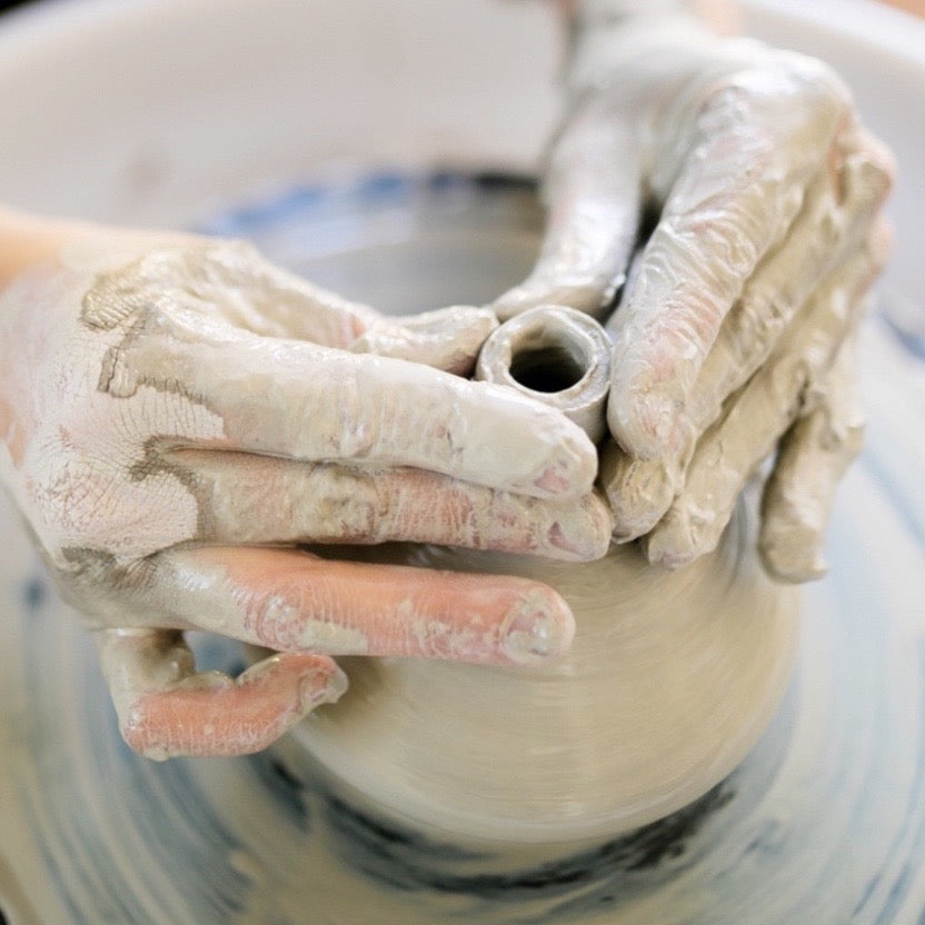 Clay-covered hands centering a ceramic vessel on a pottery wheel