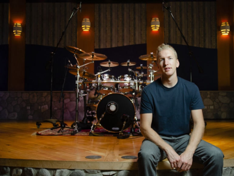 Jared Falk seated at the edge of a studio stage with a drum kit behind him
