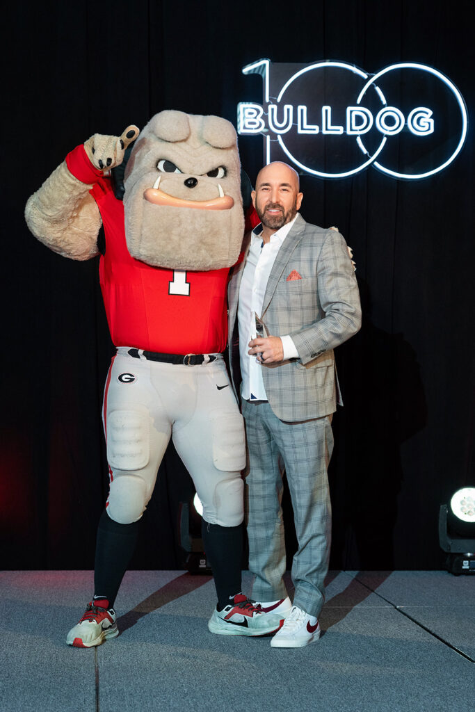 Jason Russell in a plaid suit standing beside the University of Georgia bulldog mascot at a Bulldog 100 event
