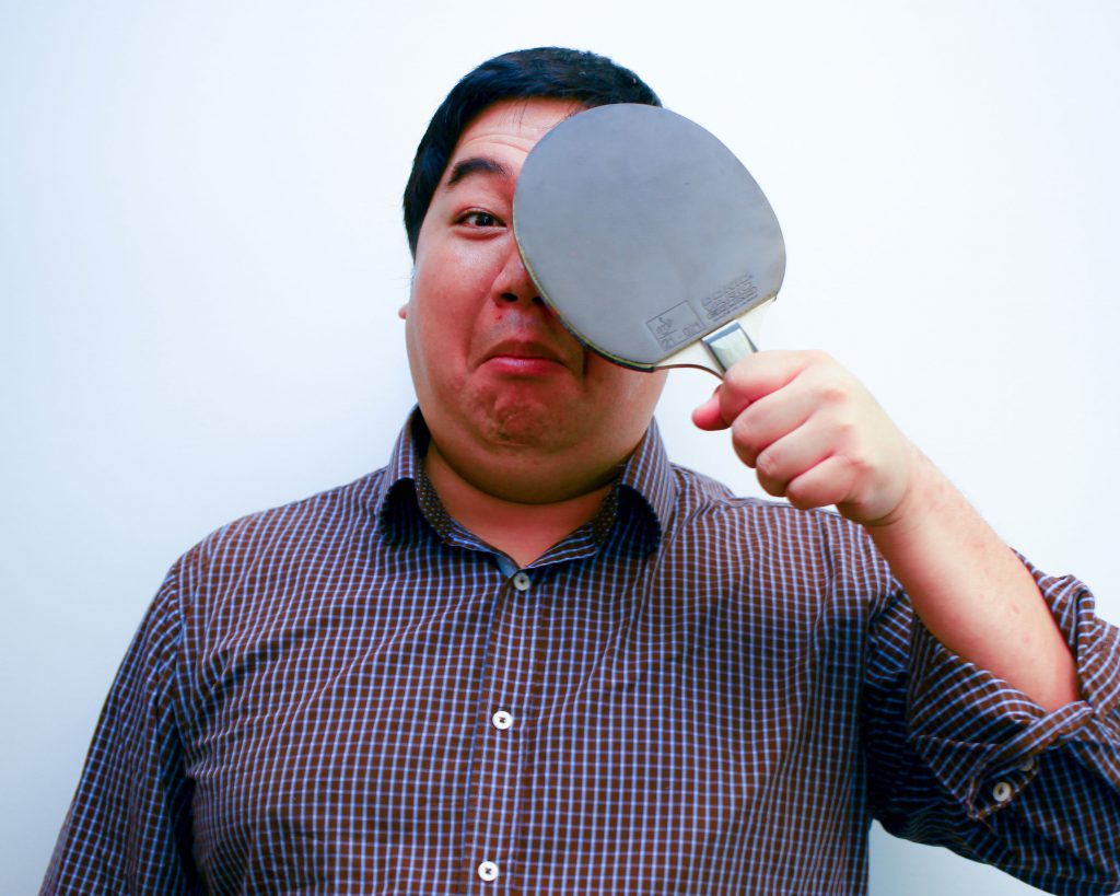 Jason Tan playfully holding a ping-pong paddle over one eye