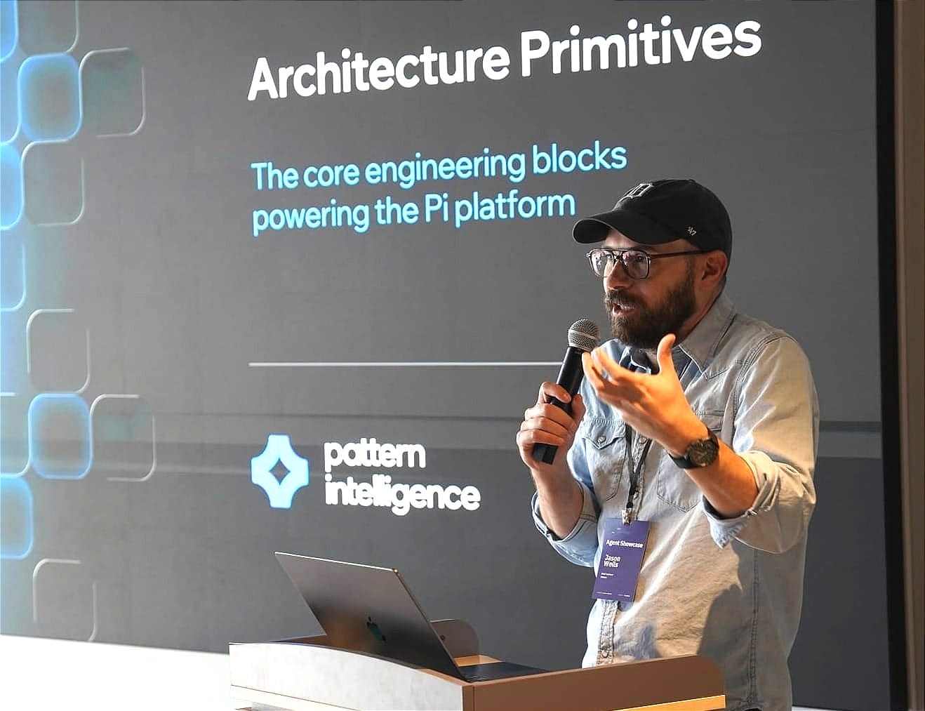 Jason Wells speaking into a microphone at Pattern’s July 2026 AI showcase, with an architecture slide behind him