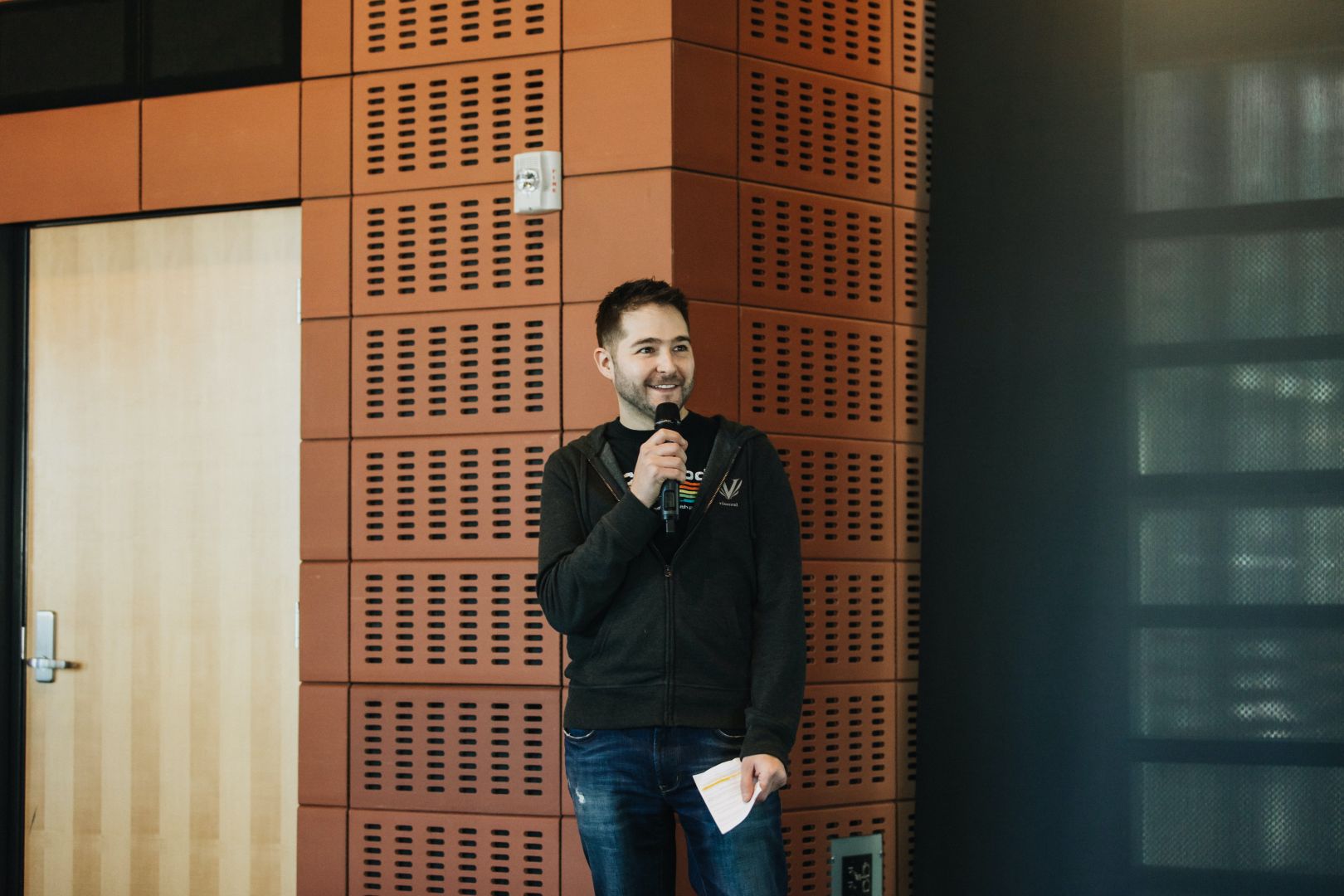 Jay Buys holding a microphone and notes while speaking in front of a wood-paneled wall