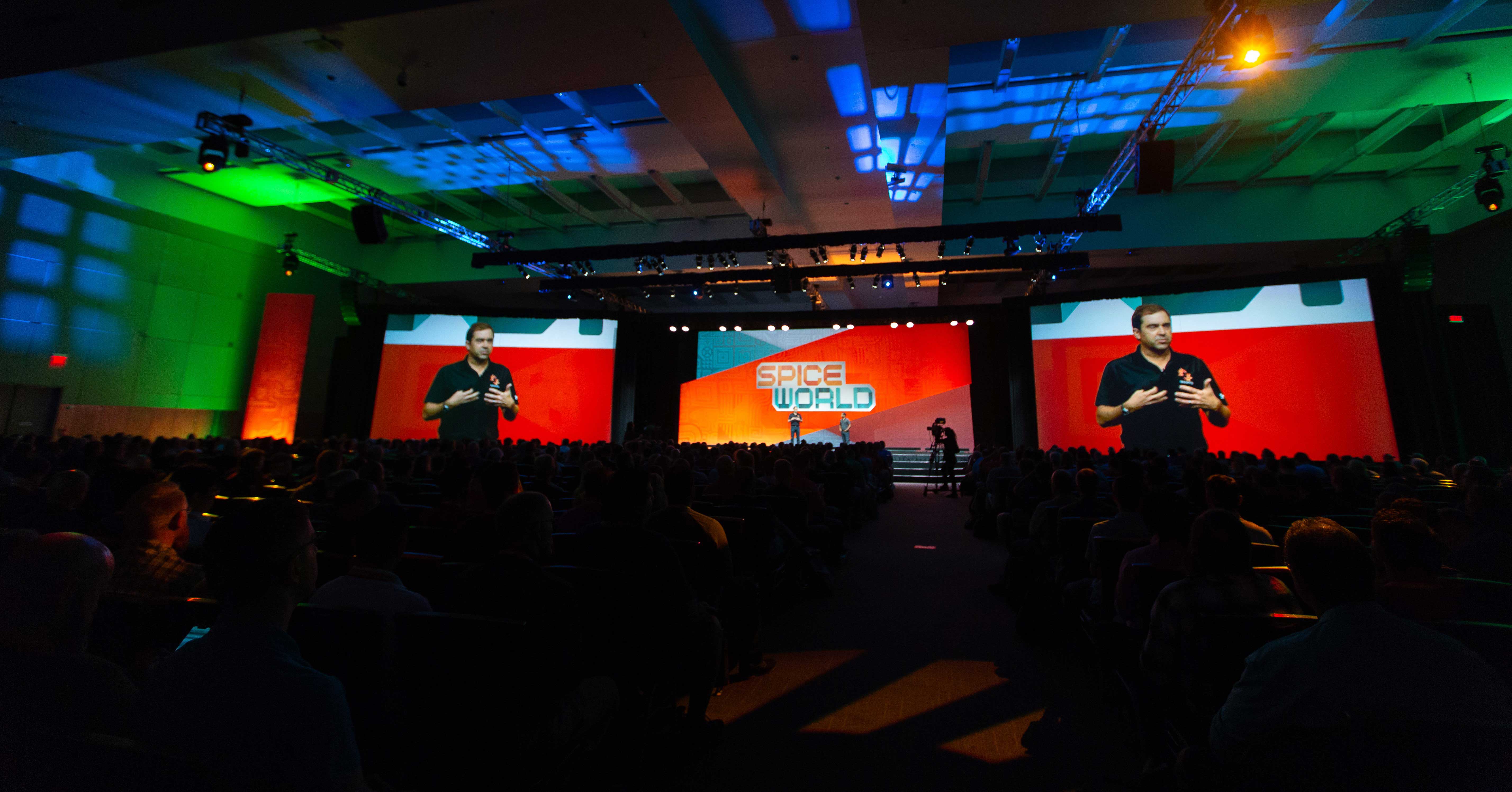 Jay Hallberg speaking on the SpiceWorld stage, with his image projected above a seated audience