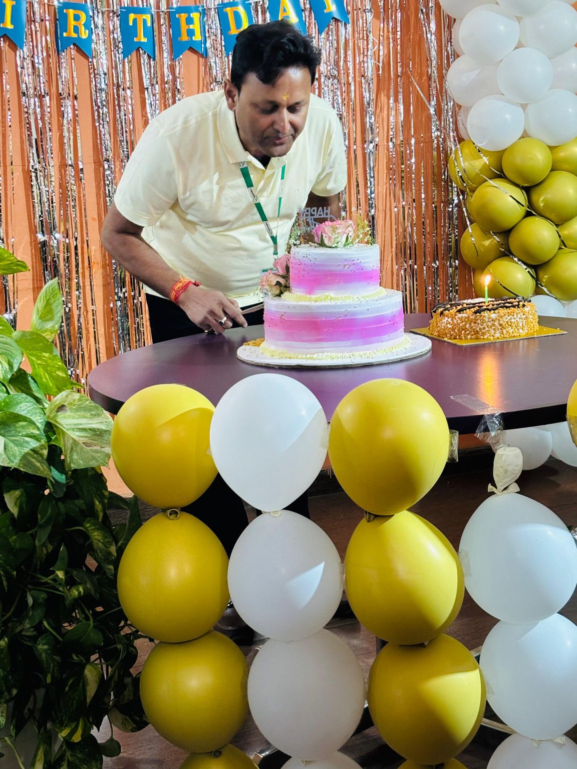 Jitendra Singh at an office birthday celebration, leaning toward a pink cake beneath white and yellow balloons.