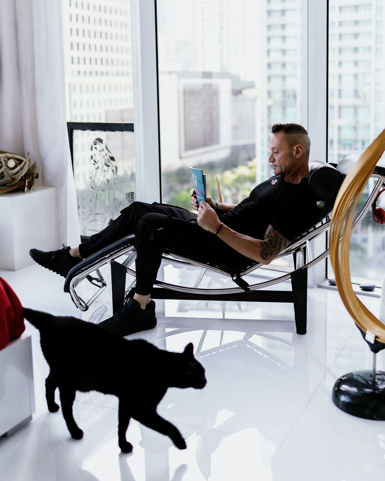 Jean Fallacara reading in a reclining chair beside large windows, with a black cat in the foreground