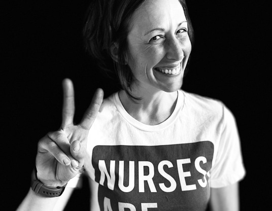 Jean Ross smiling in a nurses T-shirt and holding up two fingers