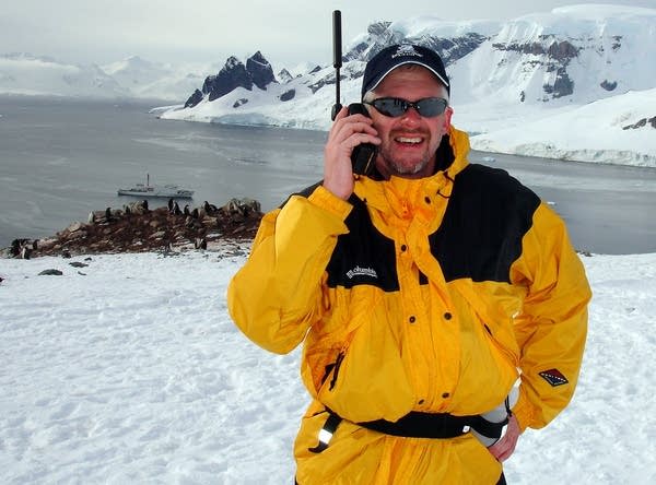 Jeff Smedsrud in a yellow jacket, holding a handset in Antarctica, with penguins and ice behind him