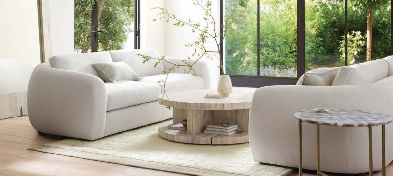 An Arhaus living room arrangement with curved white seating, pale wood tables and garden windows