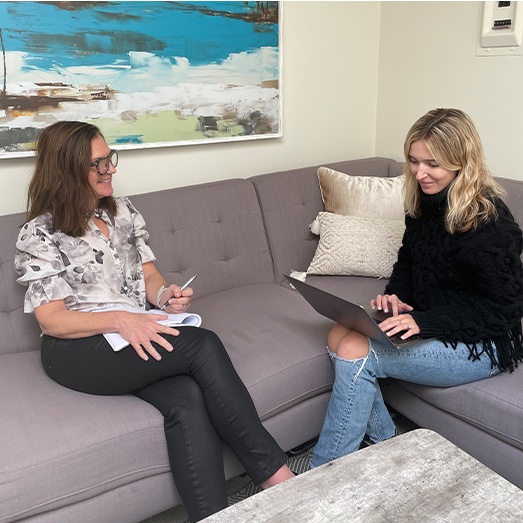 Jennifer Stephens Acree and a JSA colleague discussing work on a sofa, with a notebook and laptop.