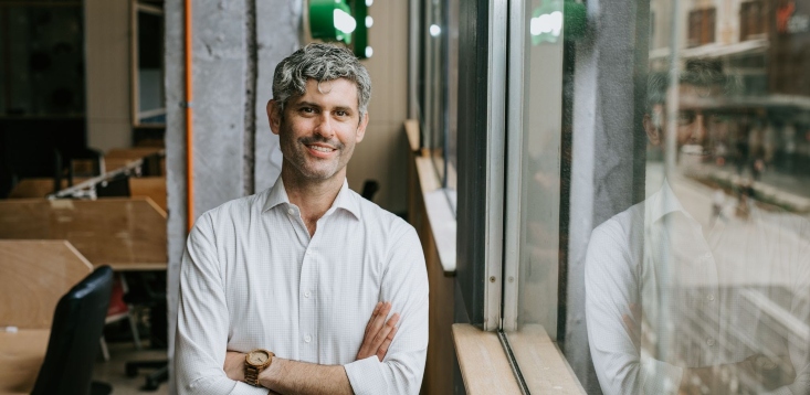 Jeremy Goldschmidt with arms folded beside a window, reflected in the glass