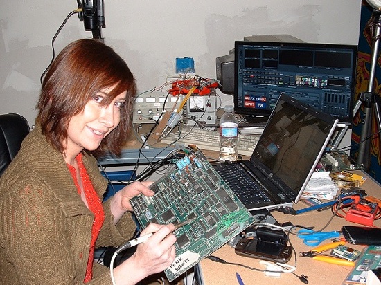 Jeri Ellsworth examining a large circuit board at a crowded electronics workbench