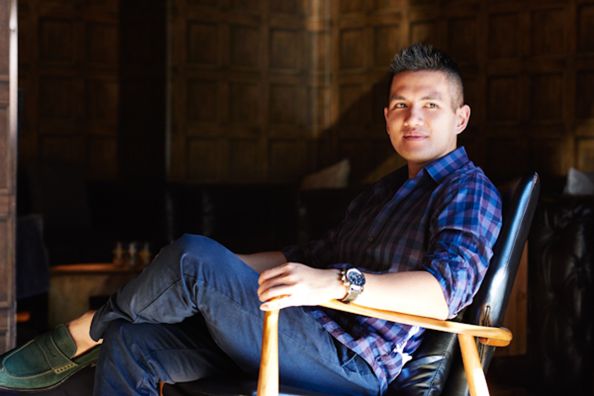 Jerry Hum seated in a wooden armchair, wearing a blue checked shirt and a wristwatch