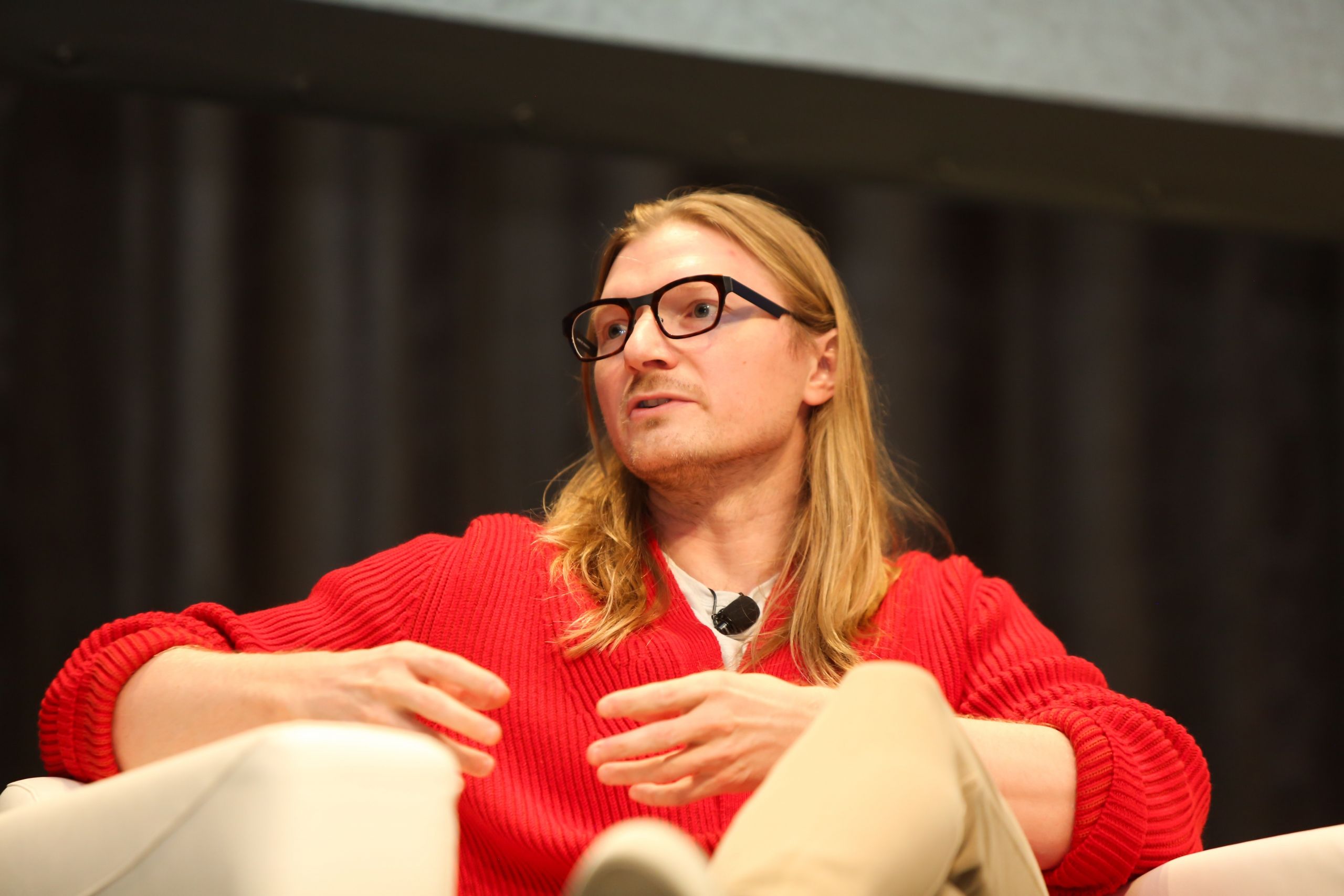 Jesse Powell speaking while seated in an armchair, wearing a red cardigan
