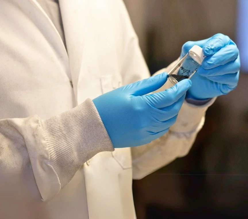 Gloved hands holding a vial of dark battery material
