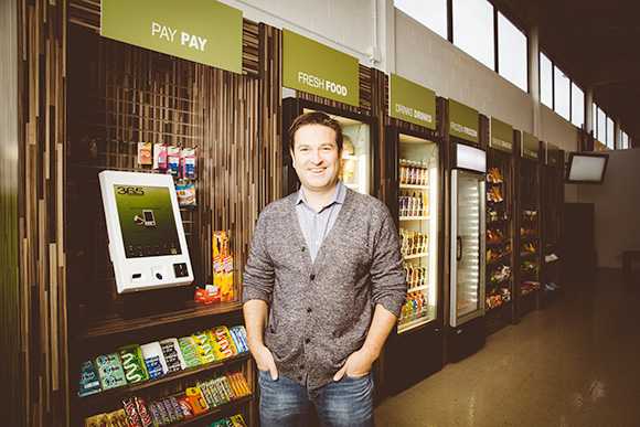 Joe Hessling standing beside a self-checkout kiosk, snack shelves and drink coolers in a workplace micro market