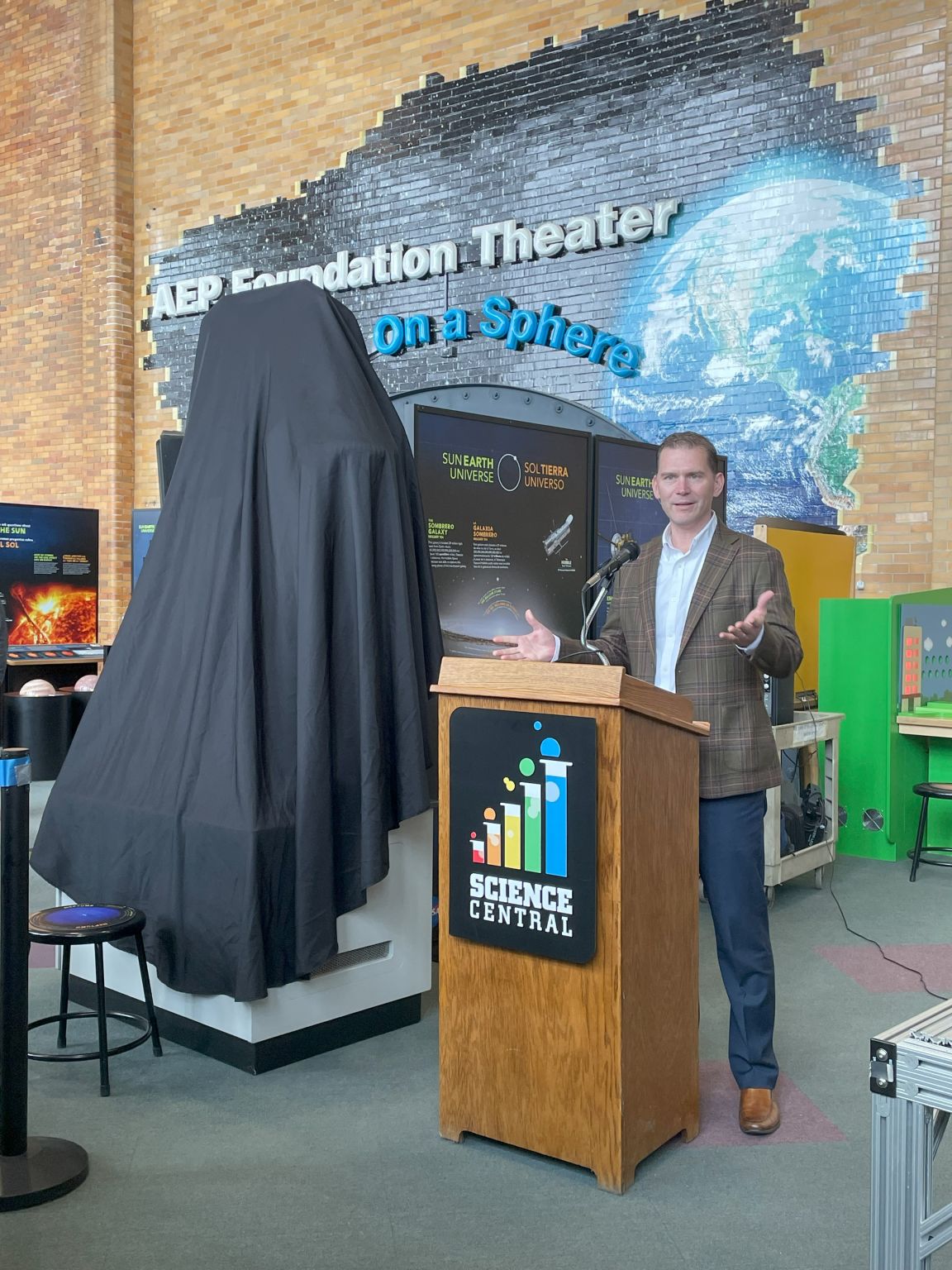 Joe Ruzynski speaking at a lectern beside a covered exhibit at Science Central in Fort Wayne