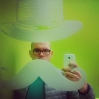John Allison taking a playful mirror selfie behind a large white moustache prop