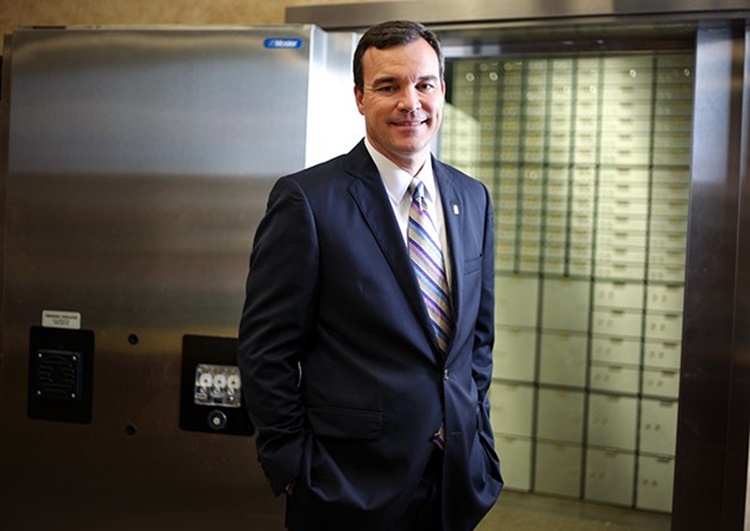 John Corbett standing beside a bank vault door and rows of safe-deposit boxes