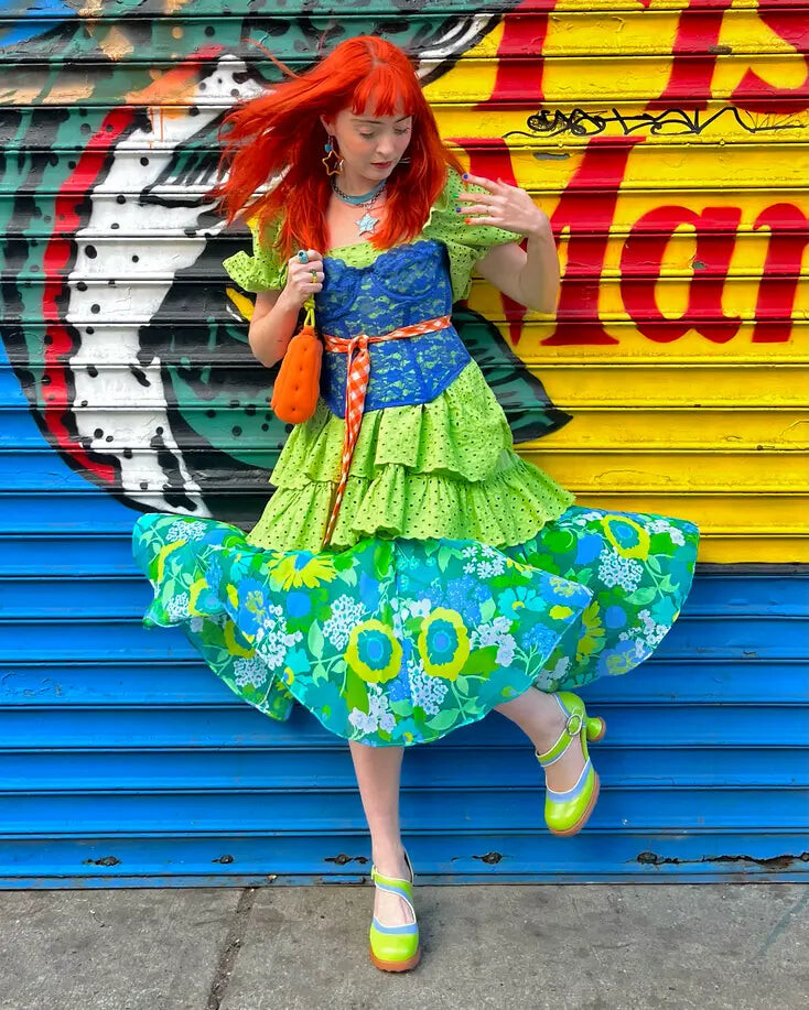 A Fluevog customer in a vivid green and blue outfit poses in front of a colorful painted shutter