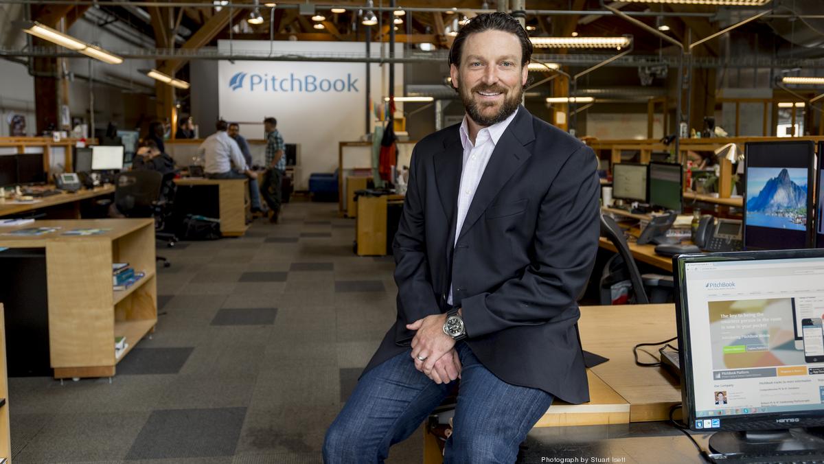 John Gabbert seated on a desk in PitchBook’s open office, with colleagues and workstations behind him.