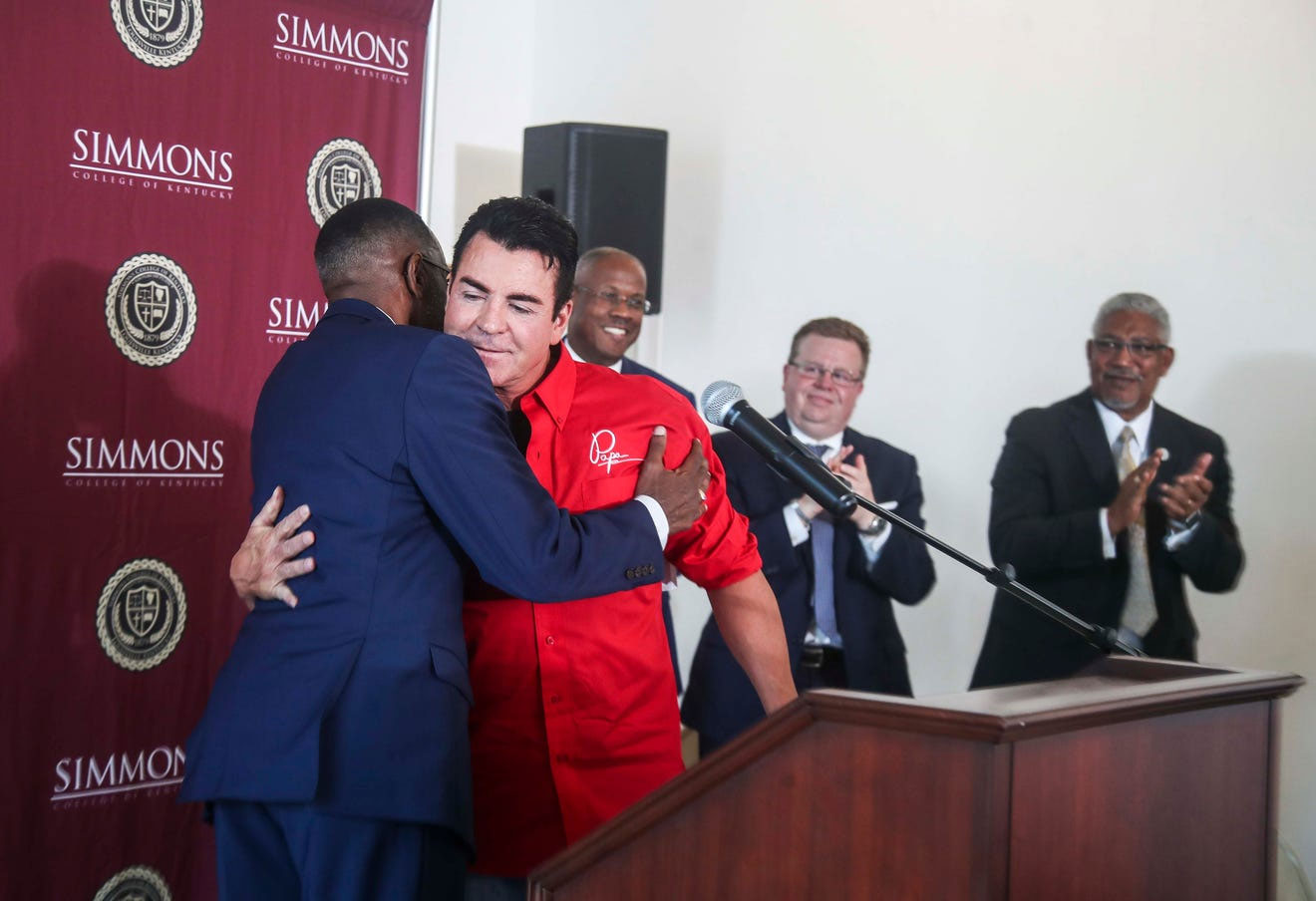 John Schnatter in a red shirt embraces a man beside a lectern at Simmons College