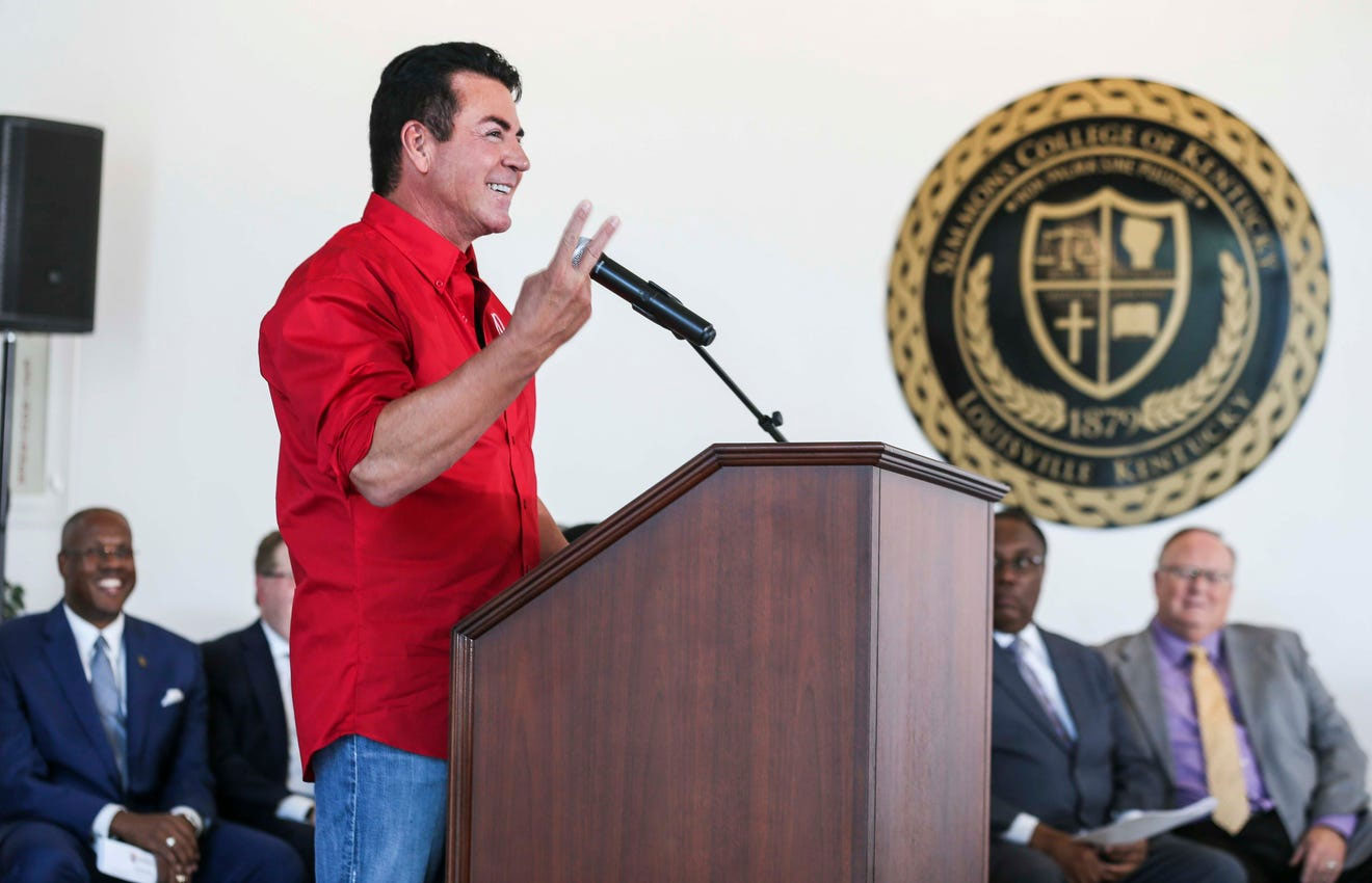 John Schnatter speaks at a lectern with Simmons College representatives seated behind him