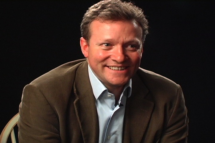 John Kearon smiling during a seated interview, wearing a brown jacket and blue shirt
