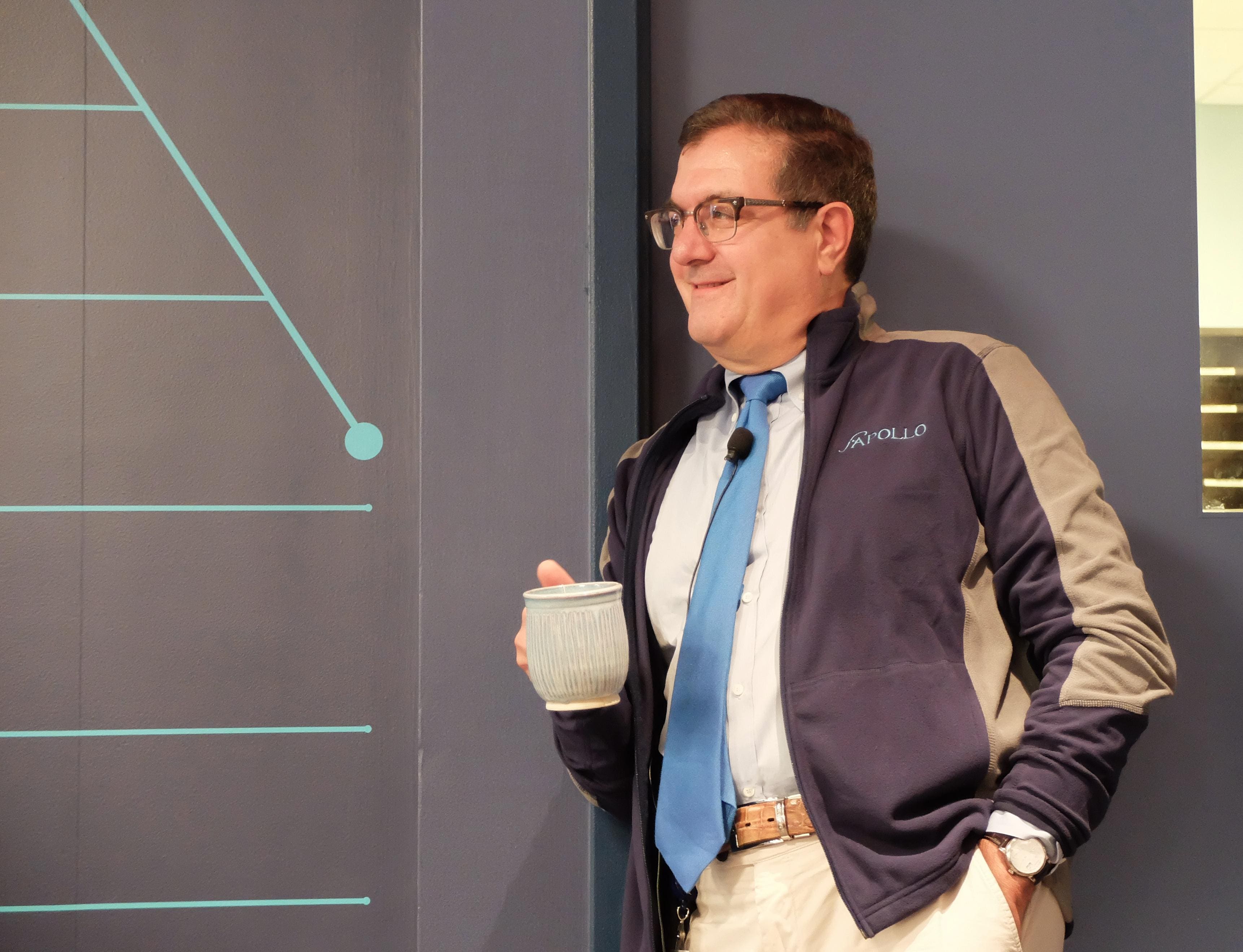 John Maraganore holding a mug and wearing a lapel microphone, an Apollo jacket, and a blue tie.