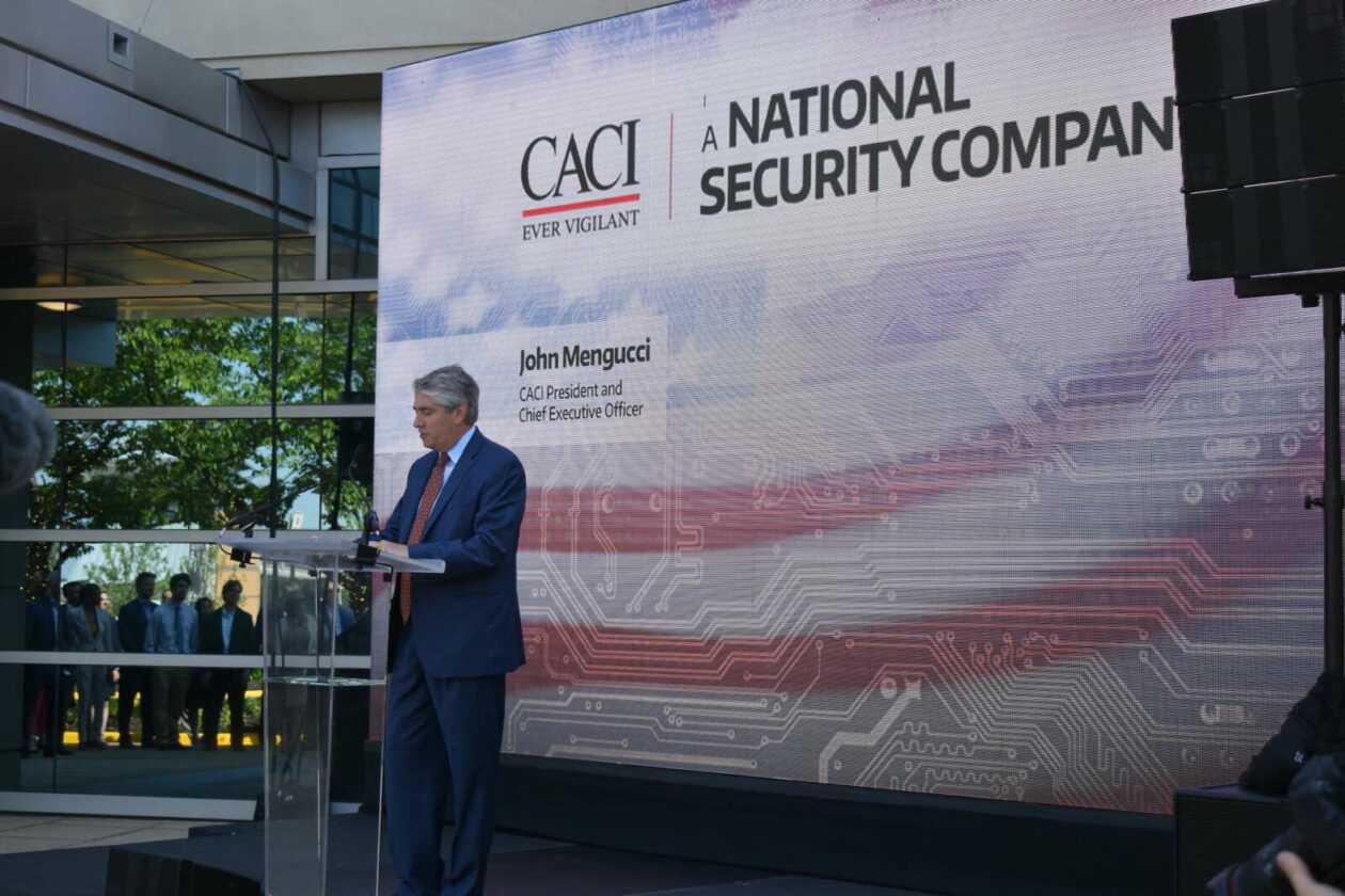 John Mengucci speaking at a lectern outside CACI’s Reston headquarters, with a large company screen behind him