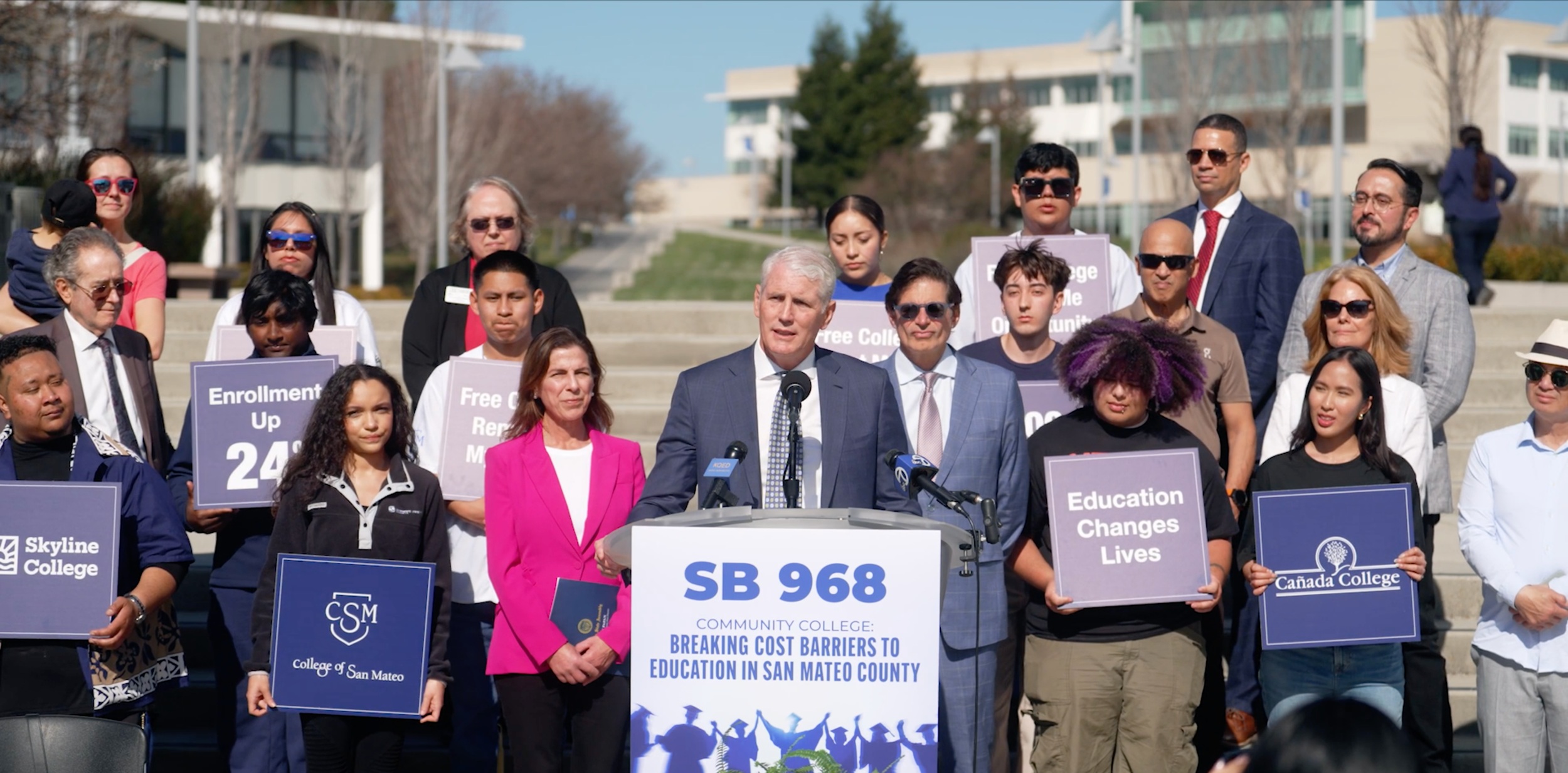 John Pimentel speaking at a podium marked SB 968, surrounded by people holding college signs